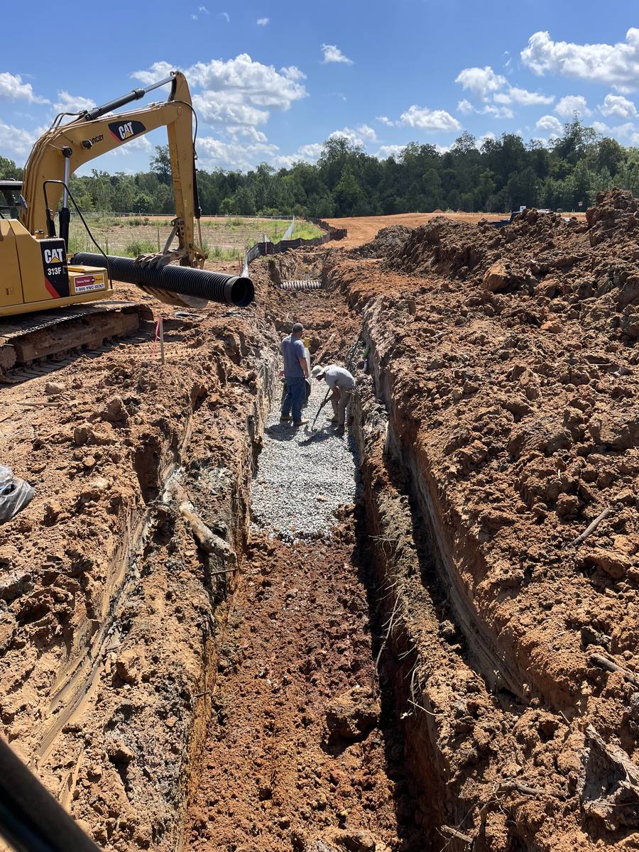 Excavator lowering pipe into a stone-bedded trench with the crew in the trench